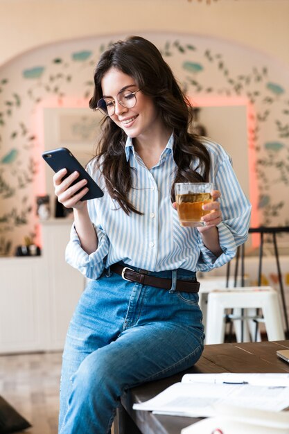 Jolie jeune femme brune souriante étudiant dans le café à l'intérieur, à l'aide d'un téléphone portable, tenant un verre avec du jus