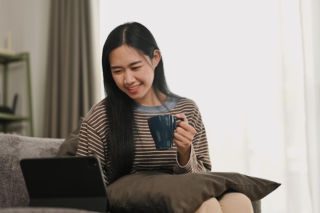 Une jolie jeune femme buvant du thé chaud et naviguant sur Internet sur une tablette numérique à la maison