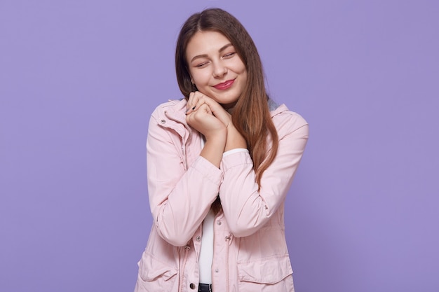 Jolie jeune femme charmante souriante vêtue d'une veste rose pâle tenant les poings près des joues isolées sur le mur lilas, gardant les yeux fermés, a l'air mignon, femme avec de longs cheveux magnifiques.