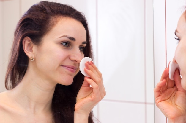 Jolie jeune femme en chemise utilisant de la crème pour le visage devant le miroir de sa salle de bain