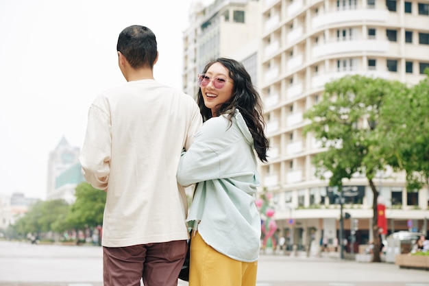 Jolie jeune femme chinoise à lunettes tenant son petit ami sous le bras se tournant vers la caméra et souriant