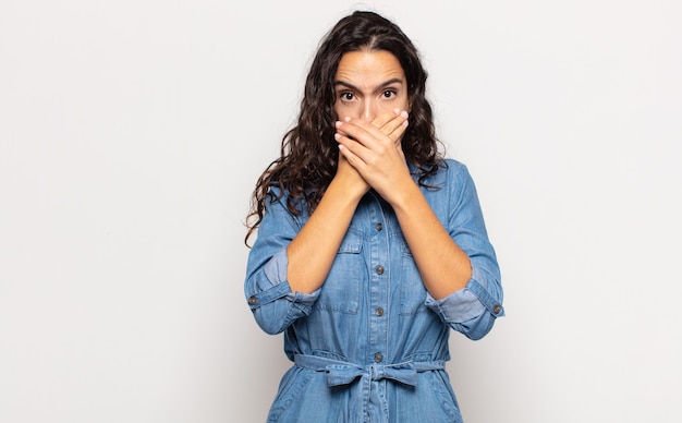 Jolie jeune femme couvrant la bouche avec les mains avec une expression choquée et surprise, gardant un secret ou disant oups