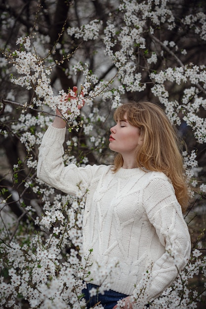 Jolie jeune femme dans un chandail blanc dans le jardin de fleurs de cerisier, printemps
