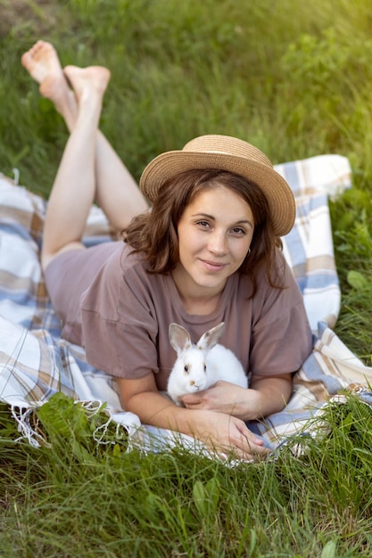 Jolie jeune femme dans un chapeau de paille se trouve sur un plaid dans un pré avec un lapin