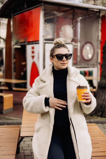 Une jolie jeune femme dans un manteau de fourrure blanche et des lunettes de soleil avec du café se promène dans la ville