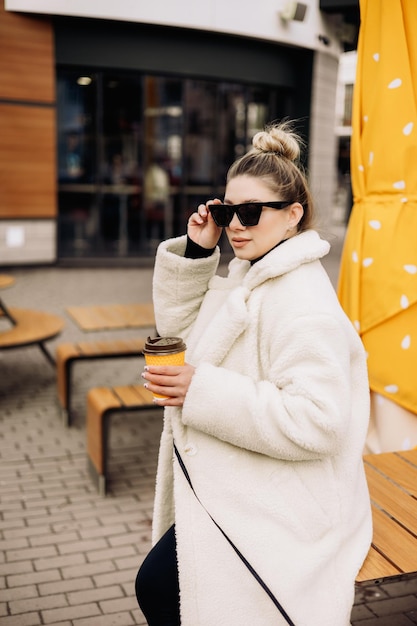 Une jolie jeune femme dans un manteau de fourrure blanche et des lunettes de soleil avec du café se promène dans la ville