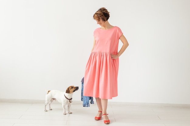 Jolie jeune femme dans une robe longue rose posant avec son chien bien-aimé sur un mur blanc. Concept de vêtements décontractés élégants. Espace publicitaire