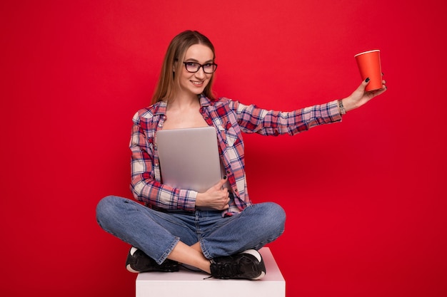 Une jolie jeune femme dans des vêtements élégants est assise avec une tasse en papier et un ordinateur portable sur fond rouge