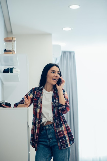 Jolie jeune femme debout dans la pièce près de la commode et souriante tout en ayant une conversation téléphonique