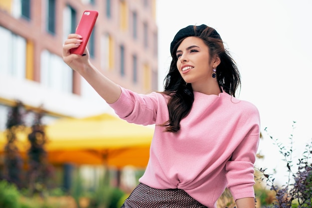 Jolie jeune femme debout à l'extérieur et prenant l'autoportrait sur un téléphone intelligent Femme brune vêtue d'un pull rose béret noir faisant selfie dans la rue