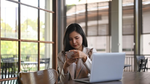 Jolie jeune femme élégante assise devant un ordinateur portable au café et utilisant un téléphone portable