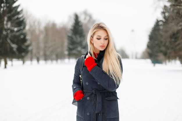 Jolie jeune femme élégante dans un manteau à la mode avec un pull rouge avec un sac élégant se promène dans le parc un jour d'hiver