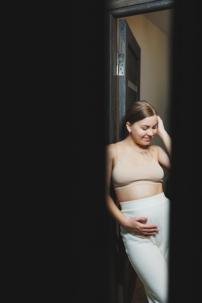Jolie jeune femme enceinte dans un haut et des vêtements de maternité décontractés Jolie femme enceinte étreignant son ventre à la maison