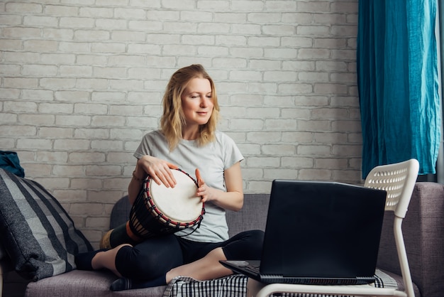 Jolie jeune femme fait de la musique en jouant du Djembé, en regardant un ordinateur portable. Cours en ligne à distance pour jouer du tambour
