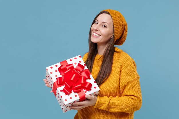 Jolie jeune femme fille en pull chapeau posant isolé sur fond bleu. Saint Valentin, Journée de la femme, anniversaire, concept de vacances. Maquette de l'espace de copie. Tenez une boîte cadeau rouge blanche avec un noeud de ruban cadeau.