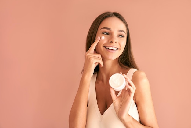 Jolie jeune femme sur fond beige tient une crème et enduit la crème sur ses joues regarde ailleurs et sourit