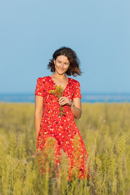 Jolie jeune femme fraîche marchant à l'extérieur dans le champ vert et vêtue d'une robe rouge d'été, tenant un bouquet de fleurs jaunes. Concept de vacances d'été au village et style de vie