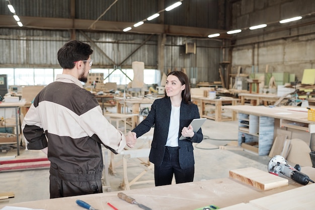 Jolie jeune femme gestionnaire ou partenaire d'affaires serrant la main d'un travailleur d'une usine de meubles après négociation dans un grand entrepôt