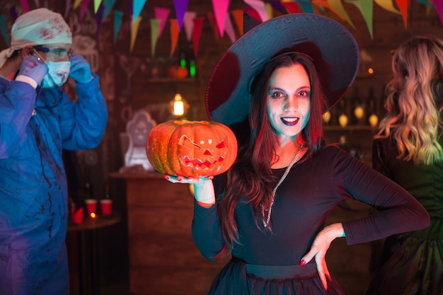 Jolie jeune femme habillée comme une sorcière pour halloween posant devant la caméra. Docteur effrayant en arrière-plan.