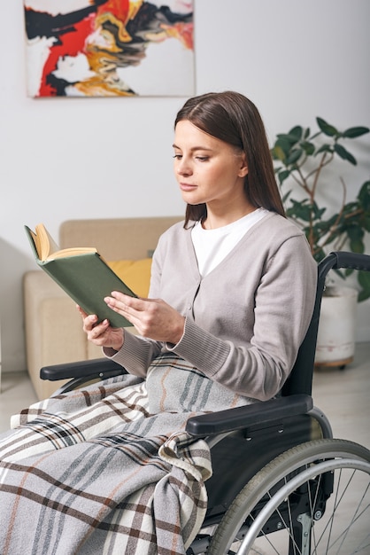 Jolie jeune femme handicapée assise sous plaid en fauteuil roulant et livre de lecture à la maison