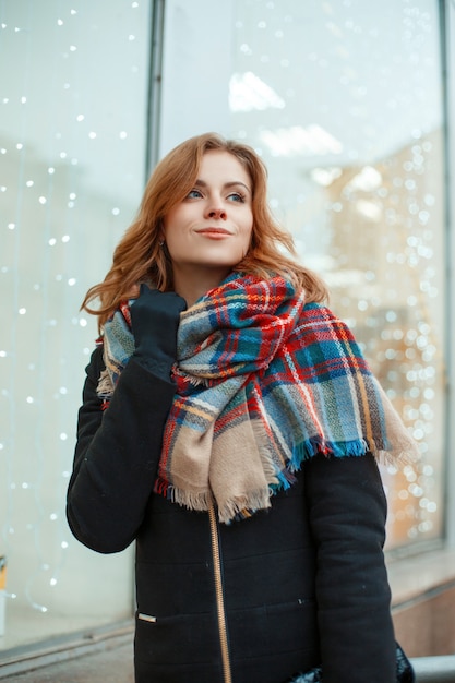 Jolie jeune femme heureuse dans un manteau d'hiver élégant en gants noirs avec une écharpe en laine se tient dans le contexte d'une vitrine décorée d'une guirlande. Joyeuse fille marchant dans la ville