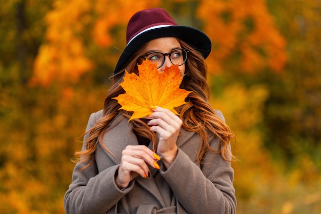 Jolie jeune femme heureuse dans des verres élégants avec un sourire positif dans un chapeau élégant dans un long manteau tient une feuille d'érable orange près du visage. Hipster fille heureuse posant avec une feuille d'automne dans le parc.