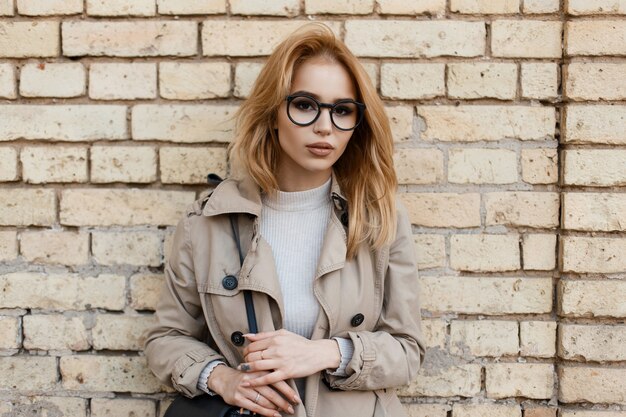 Jolie jeune femme hipster dans des lunettes élégantes en manteau tendance en T-shirt élégant blanc se tient près d'un mur de briques vintage. Charmante fille à la mode.