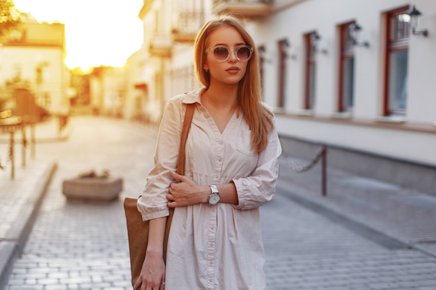 Jolie jeune femme hipster dans des lunettes de soleil élégantes dans une robe blanche élégante avec un sac à main marron à la mode se promène dans les rues vintage de la ville sur un fond de coucher de soleil. Fille à la mode