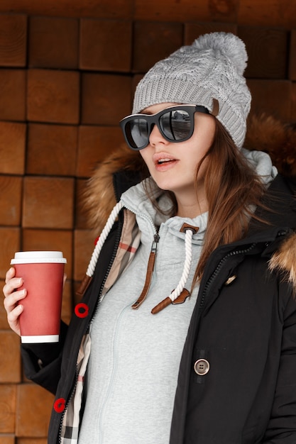 Jolie jeune femme hipster à la mode en bonnet tricoté à lunettes de soleil avec une veste noire avec une capuche en fourrure dans un sweat à capuche posant près d'un mur en bois à l'extérieur. Belle fille boit du café chaud.