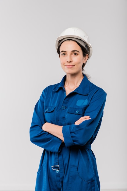 Jolie jeune femme ingénieur bras croisés en vêtements de travail bleus et casque de sécurité debout dans l'isolement