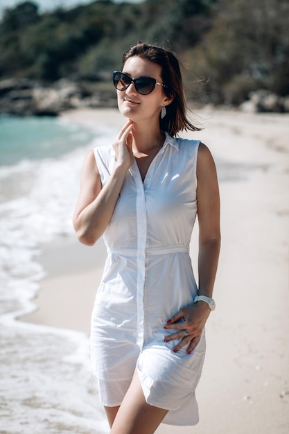 Jolie jeune femme joyeuse vêtue d'une élégante robe blanche d'été et d'une montre blanche posant sur la plage. Fille joyeuse avec un beau sourire profitez d'une journée d'été