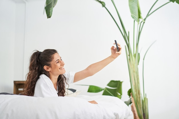 Une jolie jeune femme latina prend un selfie allongé sur le lit et partage un influenceur sur les réseaux sociaux