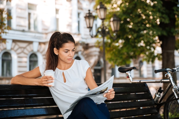 Jolie jeune femme lisant le journal alors qu'il était assis sur le banc avec une tasse de café