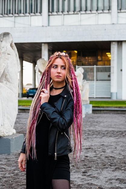 jolie jeune femme avec de longs dreadlocks roses dans une robe noire et une veste en cuir regarde pensivement sur le côté