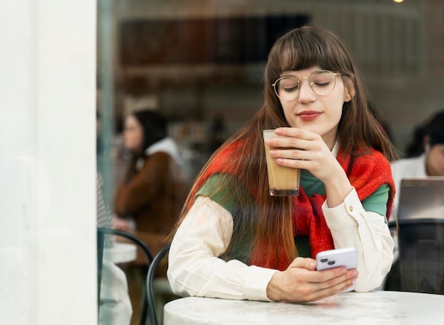 Une jolie jeune femme avec des lunettes élégantes buvant du café tenant un téléphone portable