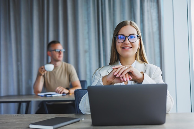 Jolie jeune femme à lunettes est assise à une table avec un ordinateur portable dans un espace de coworking et discute d'un plan de projet avec un collègue