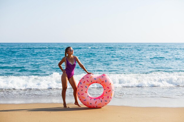 Jolie jeune femme en maillot de bain lumineux posant sur la plage avec un anneau gonflable. Belle femme blonde aux cheveux longs relaxante au bord de l'océan. Le concept d'un modèle de sport, maillots de bain