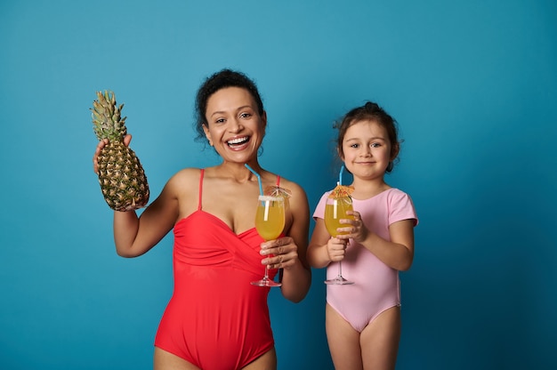 Jolie jeune femme en maillot de bain rouge tenant ananas et verre avec un délicieux jus tropical tout en se tenant à côté de son adorable fille