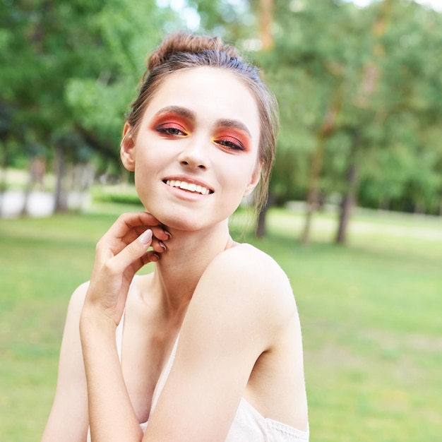 Jolie jeune femme avec un maquillage lumineux rouge et orange Outdoor green portrait park Happy smile model face Mascara style artiste Fashion smokey