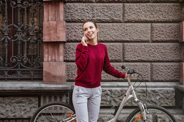 Jolie jeune femme marchant avec son vélo dans la ville tout en se tenant près du mur et en parlant par son téléphone