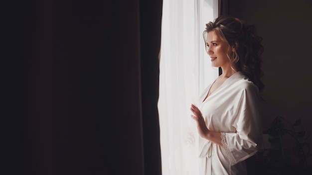 Jolie jeune femme mariée en peignoir à la maison à la fenêtre dans la chambre d'hôtel. Matin de la mariée le jour du mariage