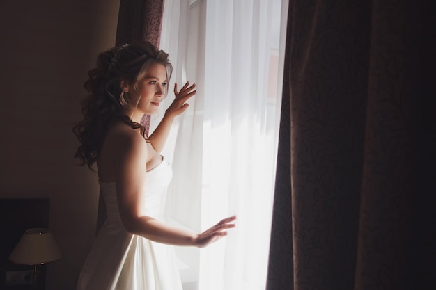 Jolie jeune femme mariée en robe blanche à la fenêtre dans la chambre d'hôtel