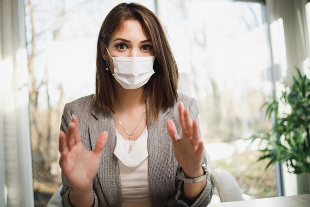 Photo une jolie jeune femme avec un masque chirurgical assise dans son bureau et parlant avec le client. regarder la caméra.