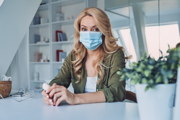 Jolie jeune femme en masque protecteur assis au bureau au bureau et regardant la caméra