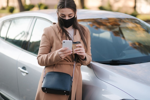 Jolie jeune femme en masque de protection avec une tasse de café près de la voiture et utiliser le téléphone.