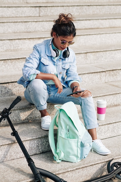 Jolie jeune femme à la mode assise sur les marches à côté de son sac à dos et vérifiant les médias sociaux sur smartphone