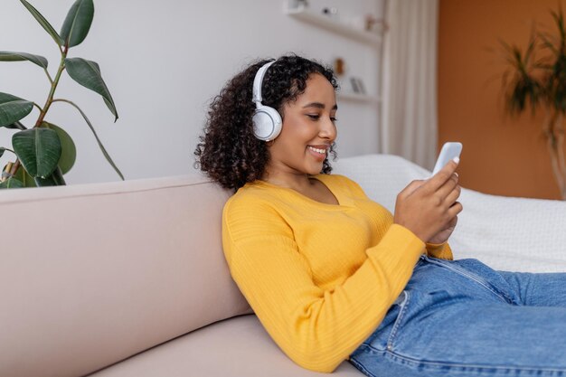 Jolie jeune femme noire dans un casque relaxant sur un canapé en écoutant de la musique ou en regardant un film sur