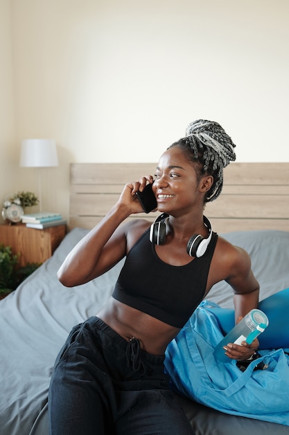 Jolie jeune femme noire en forme avec une bouteille d'eau se reposant sur le lit après l'entraînement et parlant au téléphone avec un ami