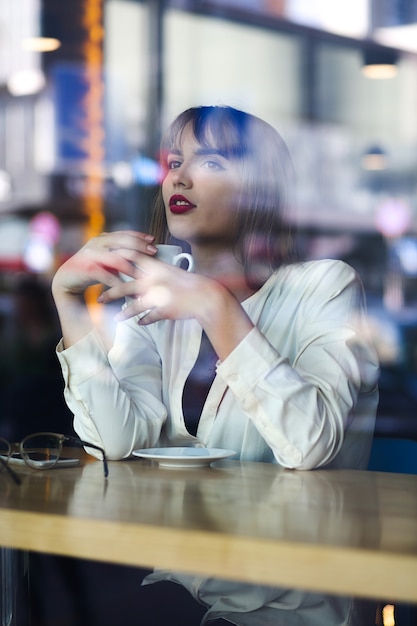 Jolie jeune femme passant du temps au restaurant avec une tasse de café. Tourné à travers une vitre