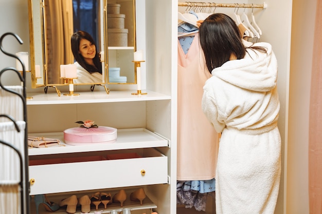 Jolie jeune femme en peignoir en choisissant une robe dans un dressing à la maison